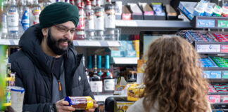 A man serves a customer at a till point inside a convenience store. The customer has placed a yellow shopping basket onto the counter with items inside it as the retailer takes items out to scan them. The retailer is holding a packet of McVitie's Chocolate Digestives.