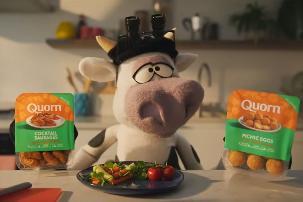 A Quorn cow puppet stands in a kitchen in front of a salad as it swaps out the meat on the plate for a meat substitute option.