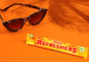 A bar of Refreshers Tropical lies on an orange floor and wall with sunglasses next to it and the shadow a palm tree behind it.