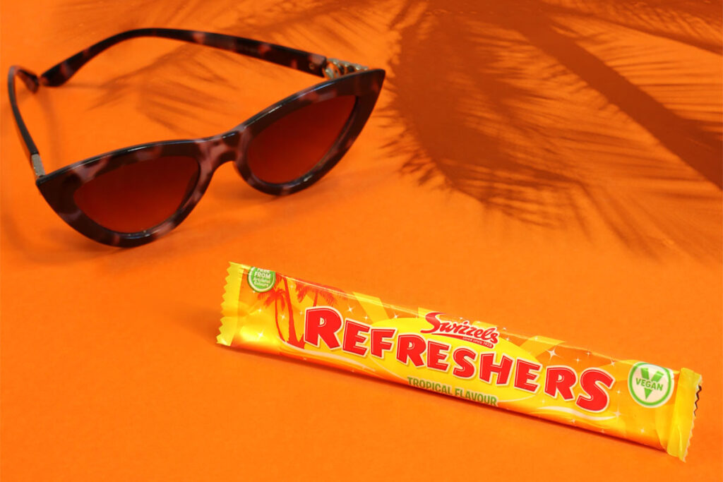 A bar of Refreshers Tropical lies on an orange floor and wall with sunglasses next to it and the shadow a palm tree behind it.