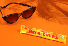 A bar of Refreshers Tropical lies on an orange floor and wall with sunglasses next to it and the shadow a palm tree behind it.