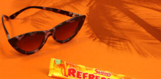 A bar of Refreshers Tropical lies on an orange floor and wall with sunglasses next to it and the shadow a palm tree behind it.