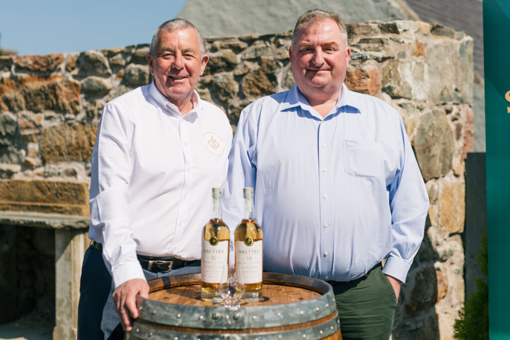 Saltire Rare Malt Founders Nigel Heywood and Keith Rennie (left to right) stand behind a whisky barrel with two whisky bottles on top of the barrel and a nosing glass in front of the whisky bottles.