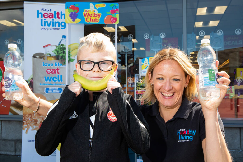 Kathryn Neil, programme director of the Healthy Living Programme, stands next to a child outside Spar Mastrick. Neil holds a bottle of water in each hand and the child holds a banana up to his mouth to look like a smile.