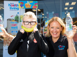 Kathryn Neil, programme director of the Healthy Living Programme, stands next to a child outside Spar Mastrick. Neil holds a bottle of water in each hand and the child holds a banana up to his mouth to look like a smile.