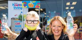 Kathryn Neil, programme director of the Healthy Living Programme, stands next to a child outside Spar Mastrick. Neil holds a bottle of water in each hand and the child holds a banana up to his mouth to look like a smile.
