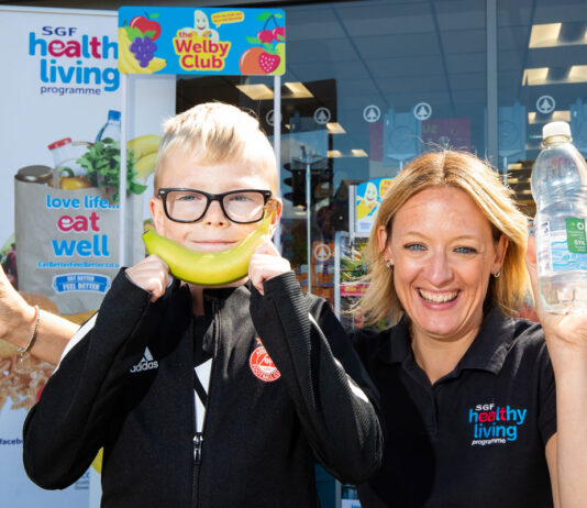 Seven Spar stores hit Premium status with Healthy Living Programme Kathryn Neil, programme director of the Healthy Living Programme, stands next to a child outside Spar Mastrick. Neil holds a bottle of water in each hand and the child holds a banana up to his mouth to look like a smile.
