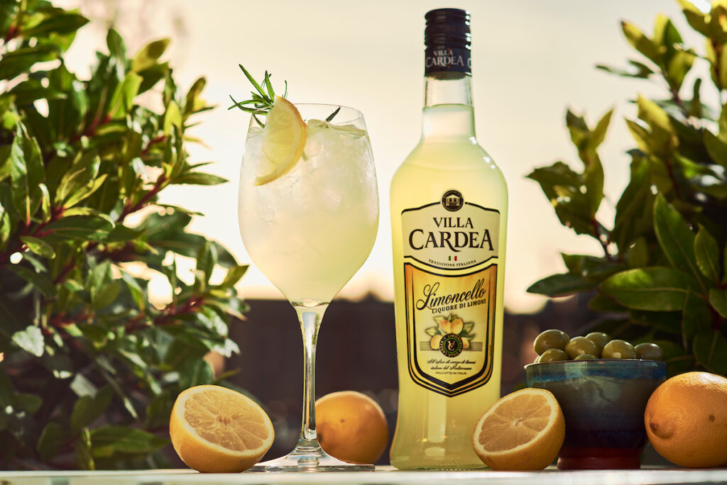 A bottle of Villa Cardea stands next to a wine glass with a Villa Cardea Limoncello Spritz cocktail in it with greenery in the background with oranges around the bottle and serve and a bowl of olives.