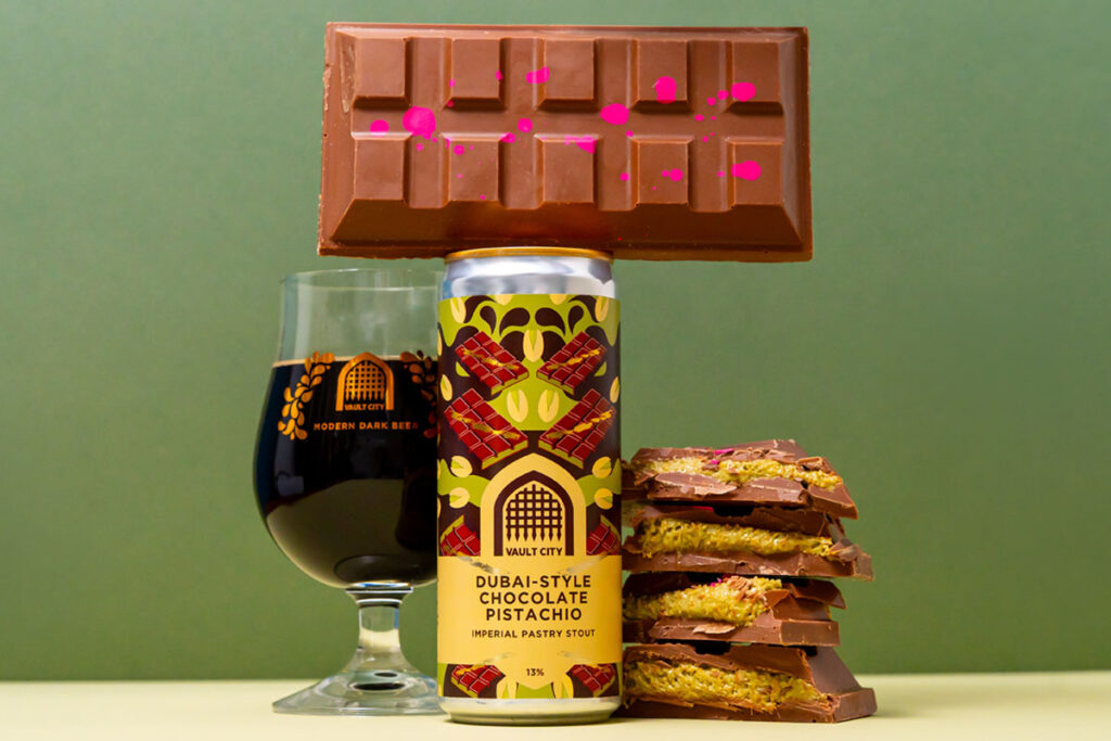 A can of Vault City Dubai Style Chocolate Pistachio Imperial Pastry Stout stands on a table with a chocolate bar balanced on top of the can, a glass with the stout inside it to the left of the image and bars of Dubai-Style Chocolate piled up to the right of the can.
