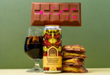 A can of Vault City Dubai Style Chocolate Pistachio Imperial Pastry Stout stands on a table with a chocolate bar balanced on top of the can, a glass with the stout inside it to the left of the image and bars of Dubai-Style Chocolate piled up to the right of the can.