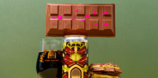 A can of Vault City Dubai Style Chocolate Pistachio Imperial Pastry Stout stands on a table with a chocolate bar balanced on top of the can, a glass with the stout inside it to the left of the image and bars of Dubai-Style Chocolate piled up to the right of the can.