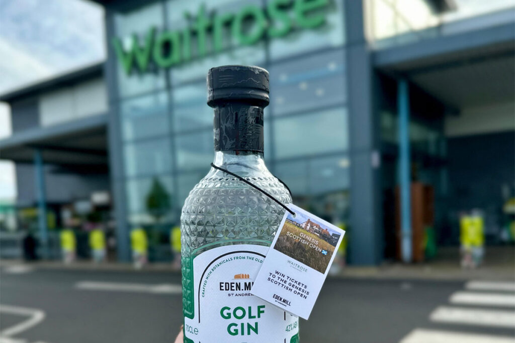 A person is holding a bottle of Eden Mill Golf Gin in front of a Waitrose supermarket store.