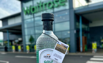 A person is holding a bottle of Eden Mill Golf Gin in front of a Waitrose supermarket store.