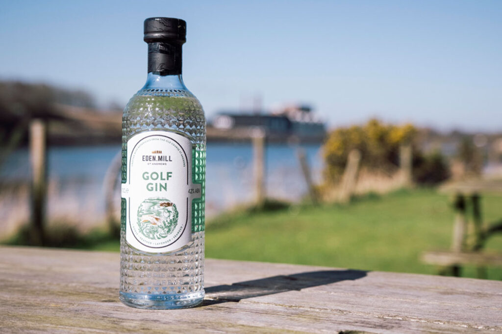 A bottle of Eden Mill Golf Gin stands on a wooden table with greenery and water in the background.