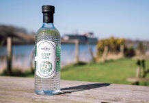 A bottle of Eden Mill Golf Gin stands on a wooden table with greenery and water in the background.