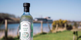 A bottle of Eden Mill Golf Gin stands on a wooden table with greenery and water in the background.