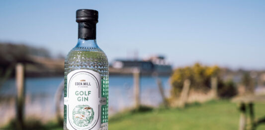 A bottle of Eden Mill Golf Gin stands on a wooden table with greenery and water in the background.