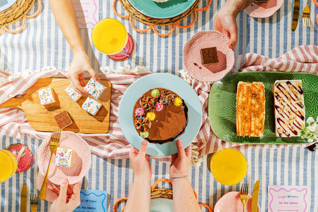 A party scene with people holding plates of cakes from the Nadiya Hussain Finsbury Food Group collection.