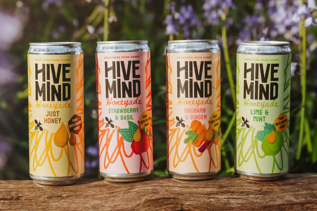 Cans of Hive Mind Honeyade drinks stand on top of a log with some greenery in the background.