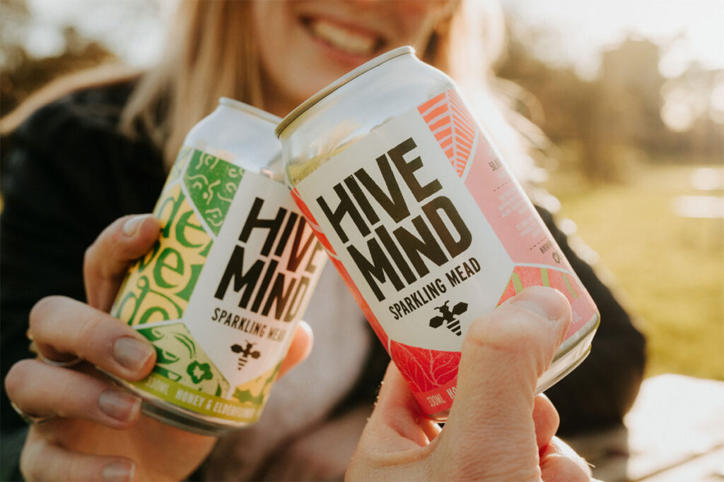 Two people clink together cans of Hive Mind Sparking Mead in a 'Cheers' action.
