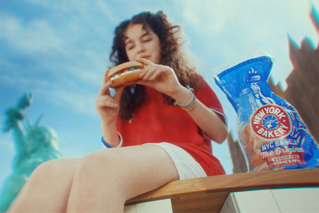 A woman sits on a bench in New York eating a bagel, there is a packet of New York Bakery Company The Original Bagels next to her and the Statue of Liberty is is the background.