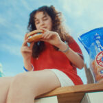 New York Bakery launches biggest marketing campaign in five years A woman sits on a bench in New York eating a bagel, there is a packet of New York Bakery Company The Original Bagels next to her and the Statue of Liberty is is the background.