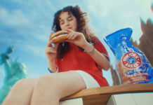 A woman sits on a bench in New York eating a bagel, there is a packet of New York Bakery Company The Original Bagels next to her and the Statue of Liberty is is the background.