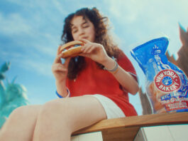 New York Bakery launches biggest marketing campaign in five years A woman sits on a bench in New York eating a bagel, there is a packet of New York Bakery Company The Original Bagels next to her and the Statue of Liberty is is the background.
