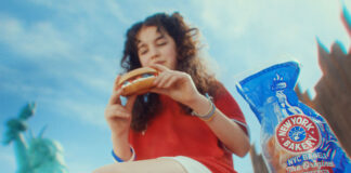 A woman sits on a bench in New York eating a bagel, there is a packet of New York Bakery Company The Original Bagels next to her and the Statue of Liberty is is the background.