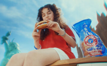 A woman sits on a bench in New York eating a bagel, there is a packet of New York Bakery Company The Original Bagels next to her and the Statue of Liberty is is the background.