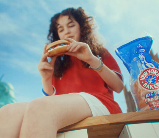 A woman sits on a bench in New York eating a bagel, there is a packet of New York Bakery Company The Original Bagels next to her and the Statue of Liberty is is the background.