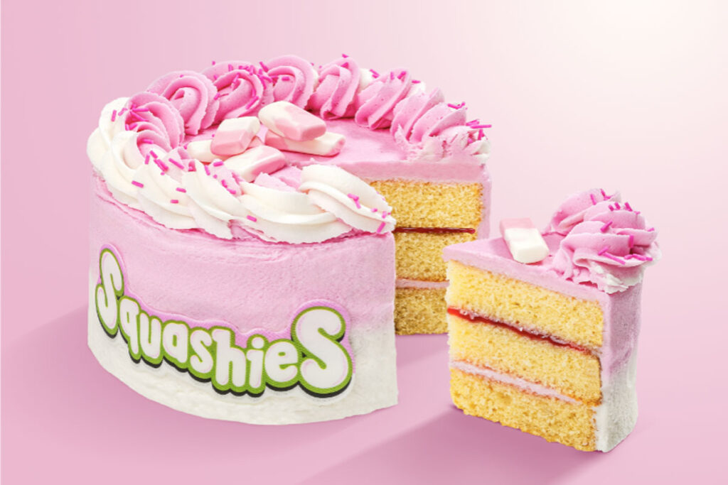A Squashies Strawberries & Cream Celebration Cake with a slice taken out of the cake against a pink background.