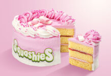 A Squashies Strawberries & Cream Celebration Cake with a slice taken out of the cake against a pink background.