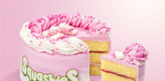 A Squashies Strawberries & Cream Celebration Cake with a slice taken out of the cake against a pink background.