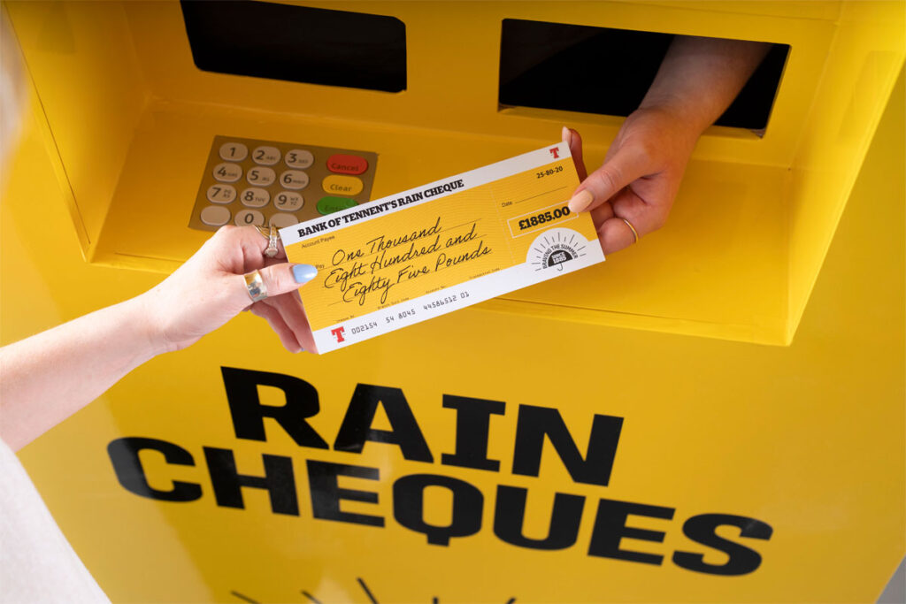 A hand passing out a cheque for £1885 with a Tennent's lager design across the cheque and the ATM machine.