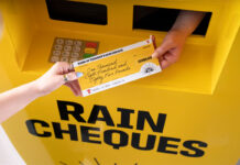 A hand passing out a cheque for £1885 with a Tennent's lager design across the cheque and the ATM machine.