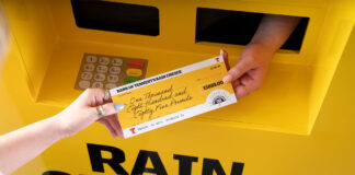 A hand passing out a cheque for £1885 with a Tennent's lager design across the cheque and the ATM machine.