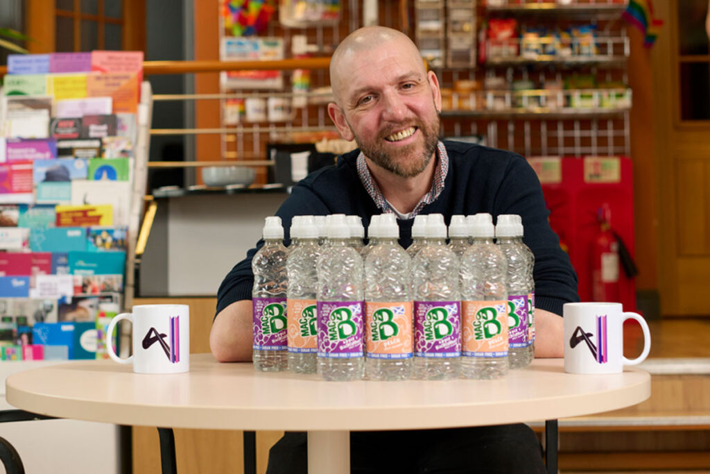 Brad Whittingham, Four Pillars LGBT+ Inclusion Advocate, sits at a table with bottles of MacB water and Four Pillars branded mugs.