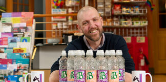 Brad Whittingham, Four Pillars LGBT+ Inclusion Advocate, sits at a table with bottles of MacB water and Four Pillars branded mugs.