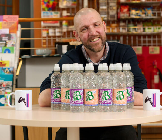 MacB brings a financial boost to Aberdeenshire charities Brad Whittingham, Four Pillars LGBT+ Inclusion Advocate, sits at a table with bottles of MacB water and Four Pillars branded mugs.