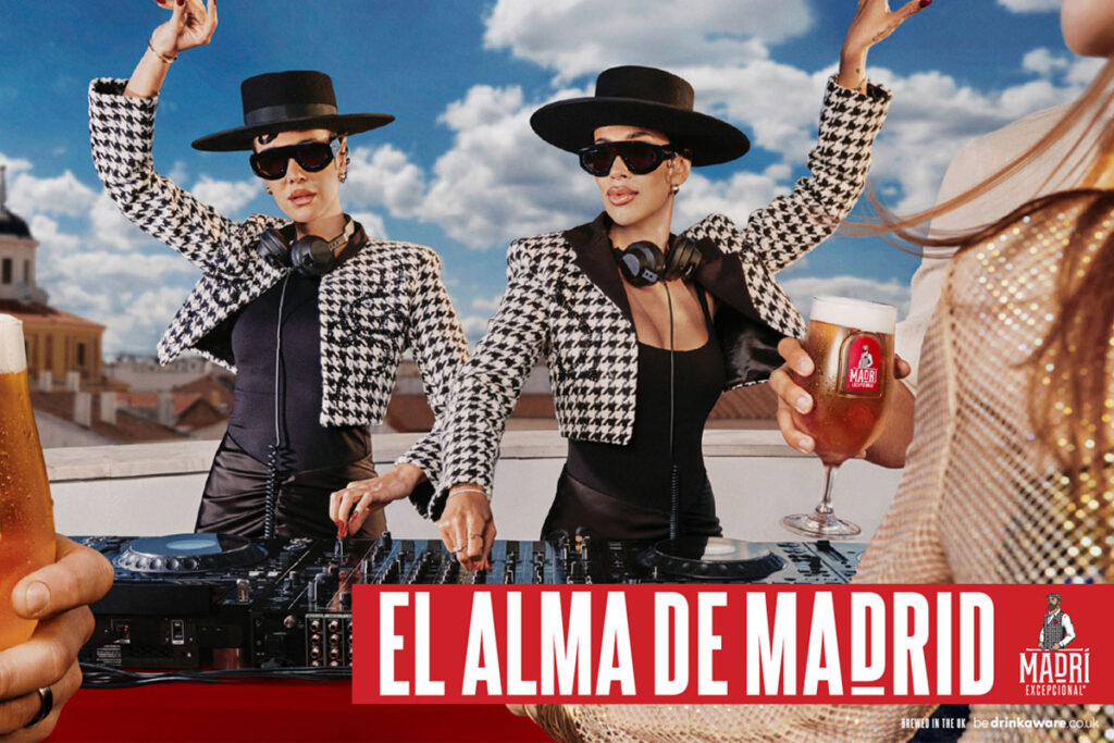 Promotional image for Madri Excepcional 2025 summer campaign with two women working on a DJ set whilst dancing, a person holds a Madri pint glass in the foreground with beer inside the glass.