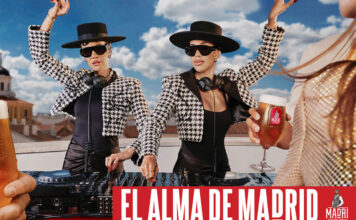 Promotional image for Madri Excepcional 2025 summer campaign with two women working on a DJ set whilst dancing, a person holds a Madri pint glass in the foreground with beer inside the glass.