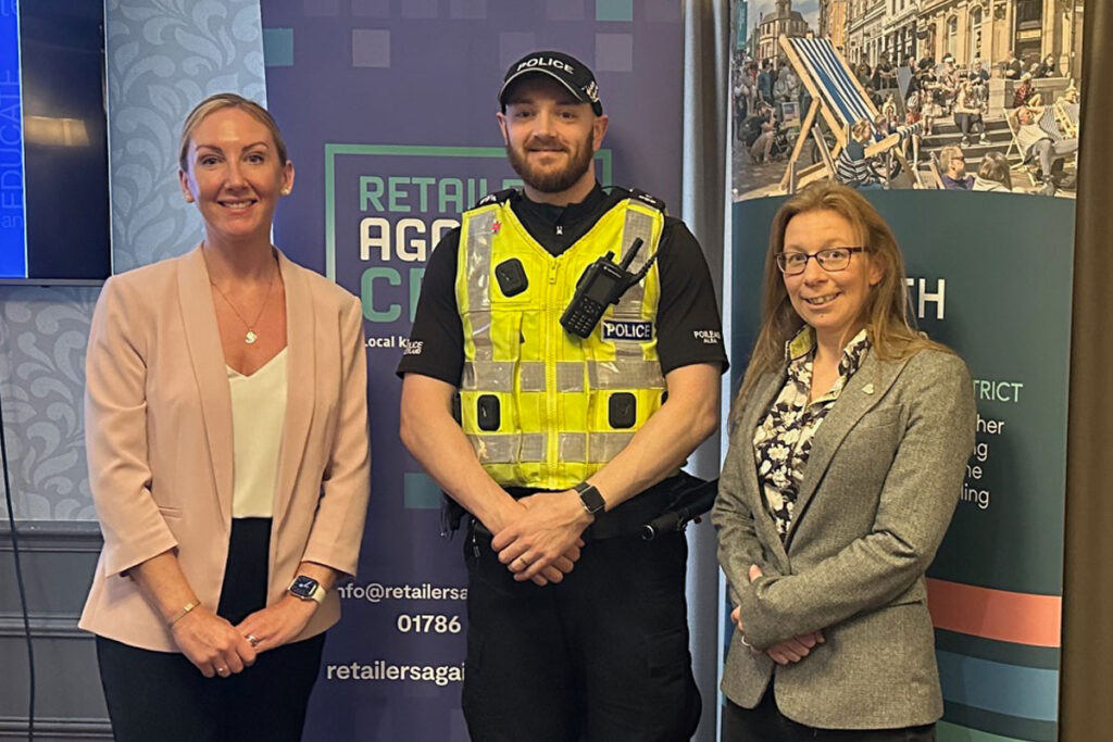 A police officer stands between two women with signage behind them for Retailers Against Crime and Go Forth Stirling.