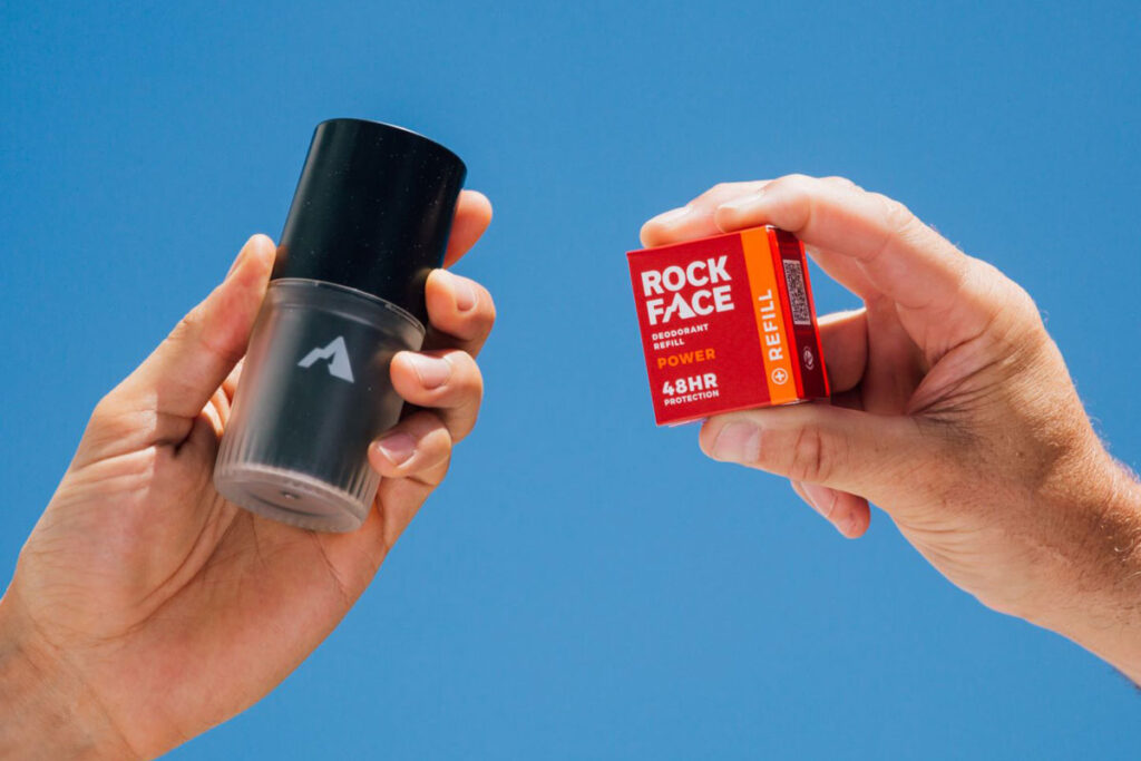 A person holds a Rock Face Deodorant Refillable container with a Rock Face Refillable Deodorant  Power pack in the other hand.