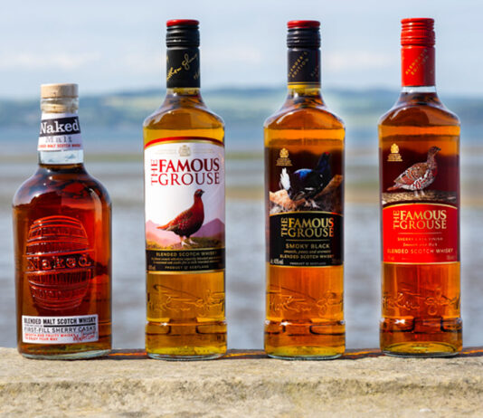 Bottle of The Famous Grouse whisky and The Naked Malt are lined up on a stone wall with an ocean view behind them.