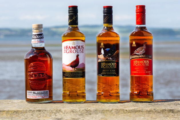Bottle of The Famous Grouse whisky and The Naked Malt are lined up on a stone wall with an ocean view behind them.