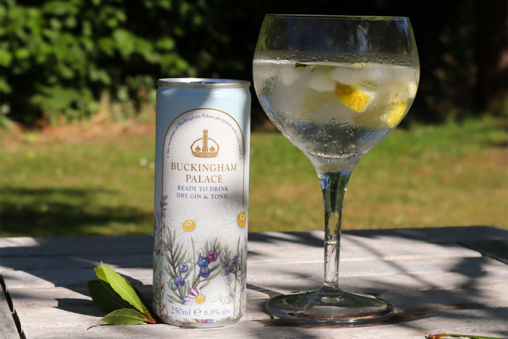 A can of Buckingham Palace Ready to Drink Dry Gin & Tonic stands next to a balloon glass with the serve inside with lemon peel and ice. The pair sit on a wooden table with greenery in the background.