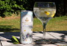 A can of Buckingham Palace Ready to Drink Dry Gin & Tonic stands next to a balloon glass with the serve inside with lemon peel and ice. The pair sit on a wooden table with greenery in the background.