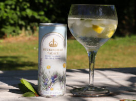 A can of Buckingham Palace Ready to Drink Dry Gin & Tonic stands next to a balloon glass with the serve inside with lemon peel and ice. The pair sit on a wooden table with greenery in the background.