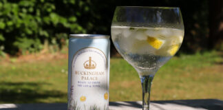 A can of Buckingham Palace Ready to Drink Dry Gin & Tonic stands next to a balloon glass with the serve inside with lemon peel and ice. The pair sit on a wooden table with greenery in the background.